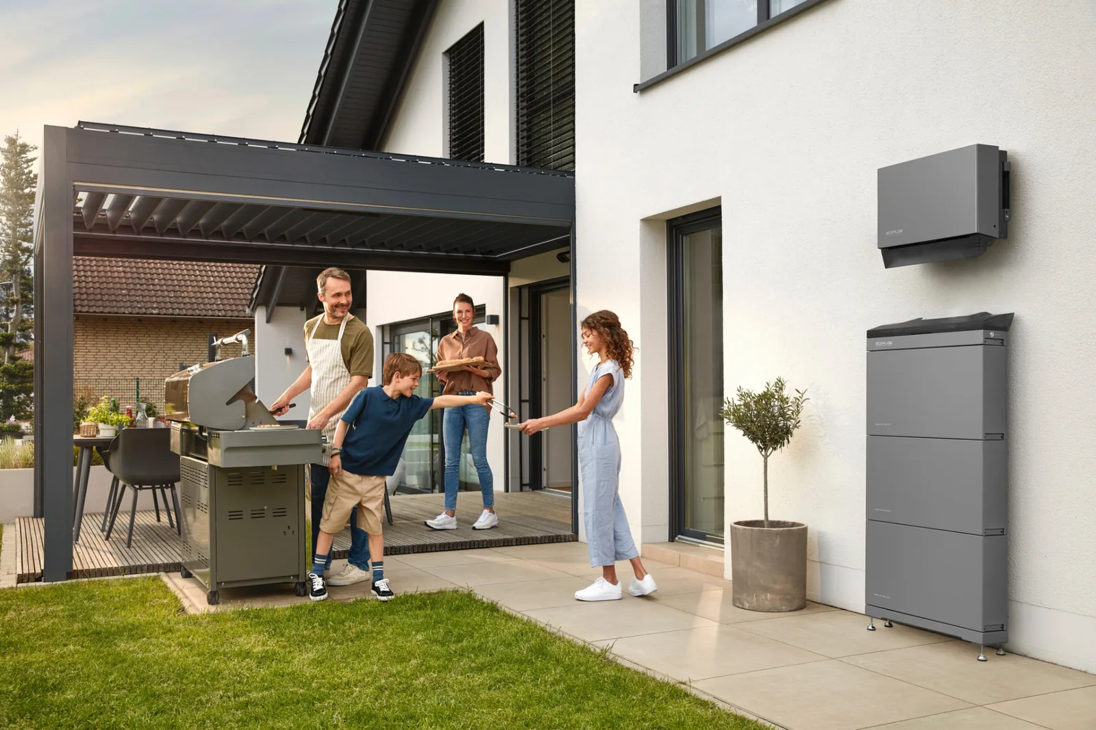 Batteriespeicher und Wechselrichter an der Außenwand eines modernen Einfamilienhauses, im Vordergrund eine Familie auf der Terrasse