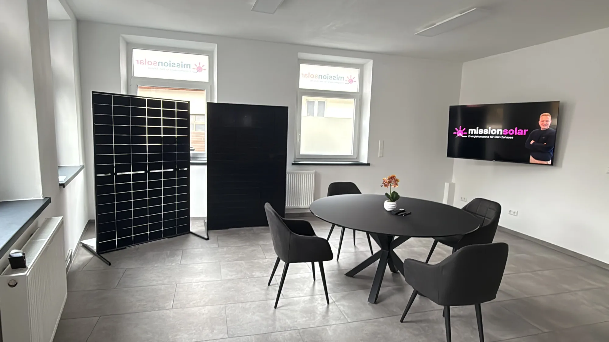 Moderner Beratungsraum mit einem runden Besprechungstisch, zwei ausgestellten PV-Modulen und einem Bildschirm an der Wand.