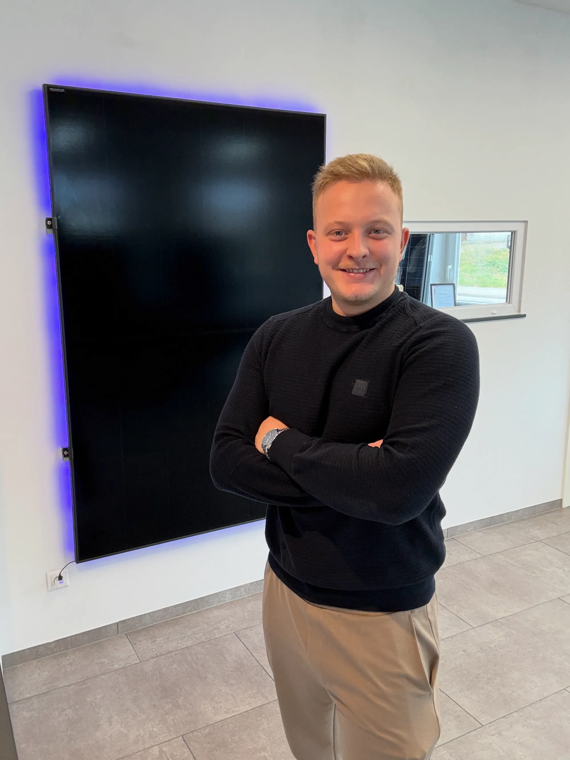 Samuel Rauscher lächelt mit verschränkten Armen vor einem blau hinterleuchteten PV-Modul.