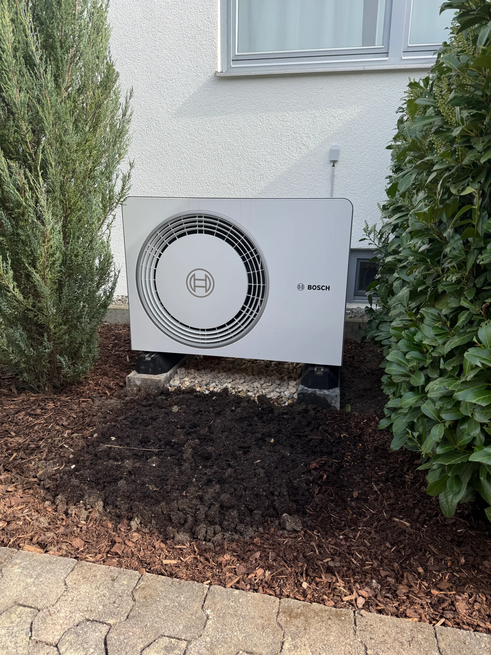 Außeneinheit einer Bosch-Wärmepumpe im Garten vor einer weiß verputzten Hauswand.